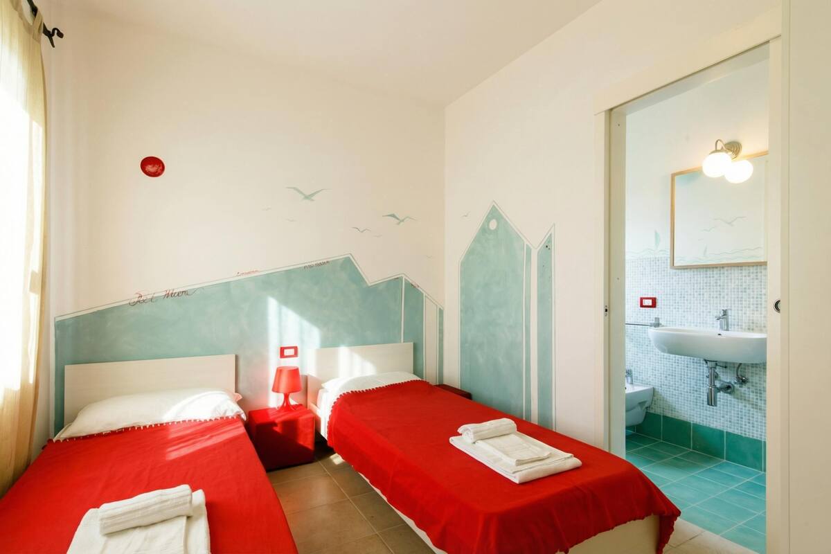 Une chambre d’hôtel avec deux lits, une décoration aux couleurs rouge et blanche, une salle de bain avec un lavabo et un miroir, et une fresque murale.