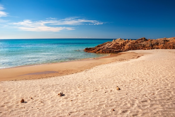 Une plage de sable fin, avec une eau d’un bleu limpide et des affleurements rocheux.