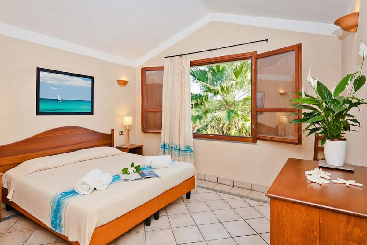 Une chambre d’hôtel avec un lit, deux tables de chevet, un bureau, une plante en pot et une fenêtre avec des rideaux.