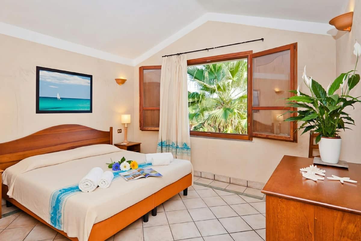 Une chambre d’hôtel avec un lit, des tables de chevet, un bureau, une plante en pot et une fenêtre donnant sur des palmiers.