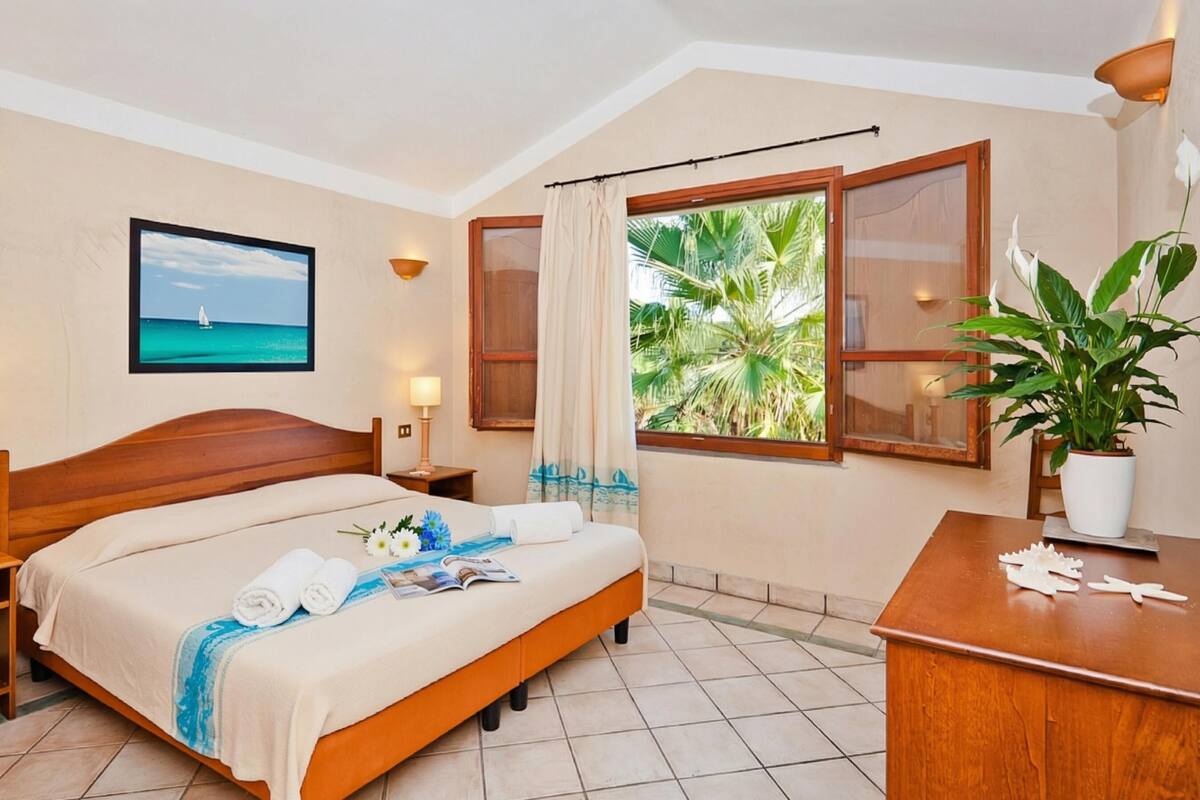 Une chambre d’hôtel avec un lit, un bureau, une fenêtre donnant sur des palmiers, un tableau encadré au mur et une plante en pot.