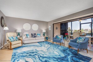 Ein geräumiges Wohnzimmer mit einem großen, blau-weißen Teppich, einem beigefarbenen Sofa, zwei Sesseln und einem Couchtisch. An den Wänden befinden sich Kunstwerke und eine Uhr.