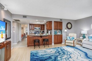 A modern kitchen with wooden cabinets, a central island with stools, and a dining area with a white sofa and a blue rug. - Intracoastal Views, Boat Dock, Beach Access, Pool (Sarasota)