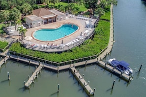 Luftaufnahme eines Resorts mit einem zentralen Schwimmbad, umgeben von Liegestühlen und einem Steg mit einem Boot.