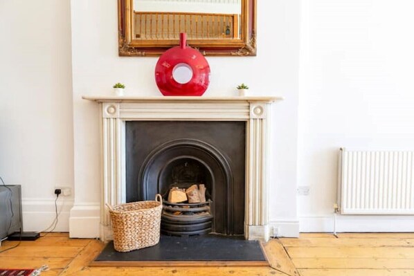 Une cheminée avec un manteau de couleur blanche, un objet décoratif rouge placé au-dessus, et un panier de bois de chauffage à côté.