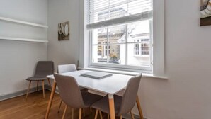 Een moderne kamer met een witte tafel en vier stoelen, een raam met jaloezieën en uitzicht op gebouwen.