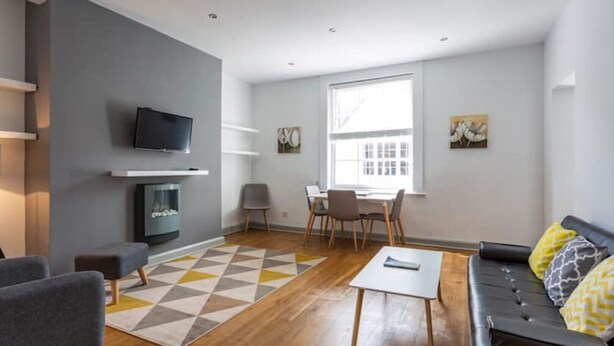 Een moderne woonkamer met een flatscreen televisie, een open haard, een eethoek met stoelen en een bank met decoratieve kussens.