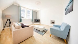 A modern living room with a sofa, a blue armchair, a coffee table, a floor lamp, and a TV. - Clifton Spacious 3 Bed Apt&Parking-Simply Check In (Bristol)