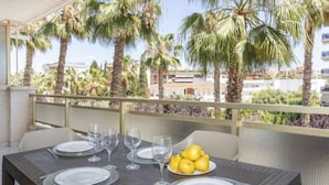 Mesa preparada para comer con platos, vasos y un cuenco de limones en un balcón con palmeras y edificios al fondo.