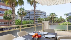 An outdoor dining area with a table set for four, surrounded by chairs. There are plates, cutlery, and glasses on the table, along with a bowl of fruit. The setting includes palm trees and a view of a building in the background. - Jardines Family 204 by Interhome (Salou)