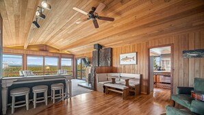 A spacious room with wooden floors, a bar area with stools, a fireplace, and a large window offering a mountain view.