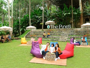 Un espace extérieur aménagé avec des fauteuils-pouf colorés et des tables, entouré d’une végétation luxuriante et d’un mur de pierre sur lequel est affiché un panneau indiquant « The Pines ».