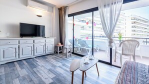 Een moderne hotelkamer met een flatscreen televisie, een witte kast, een eettafel met stoelen, en een balkon met uitzicht op de stad.