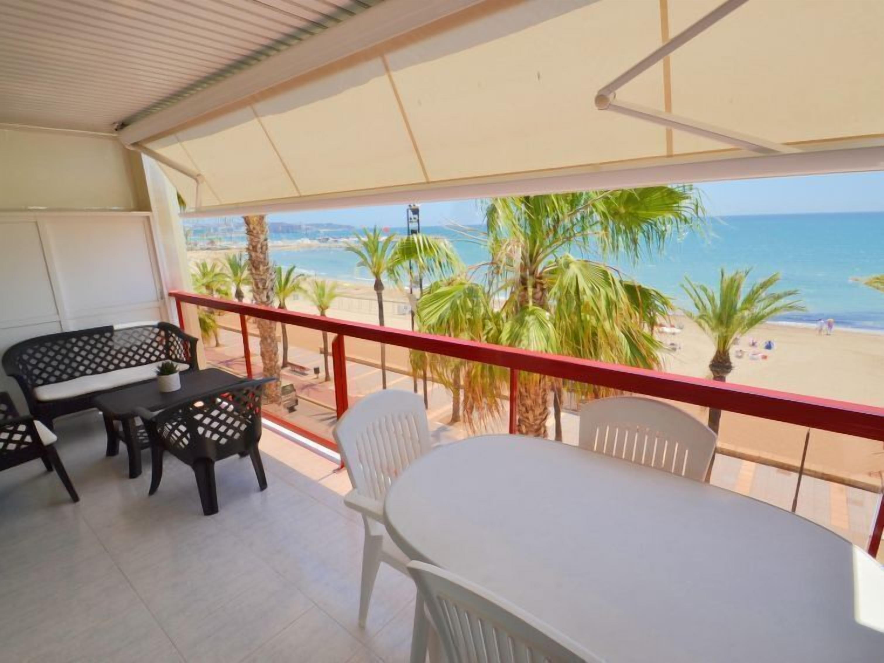 Un balcón con mesa y sillas, con vista a una playa con palmeras y el mar.