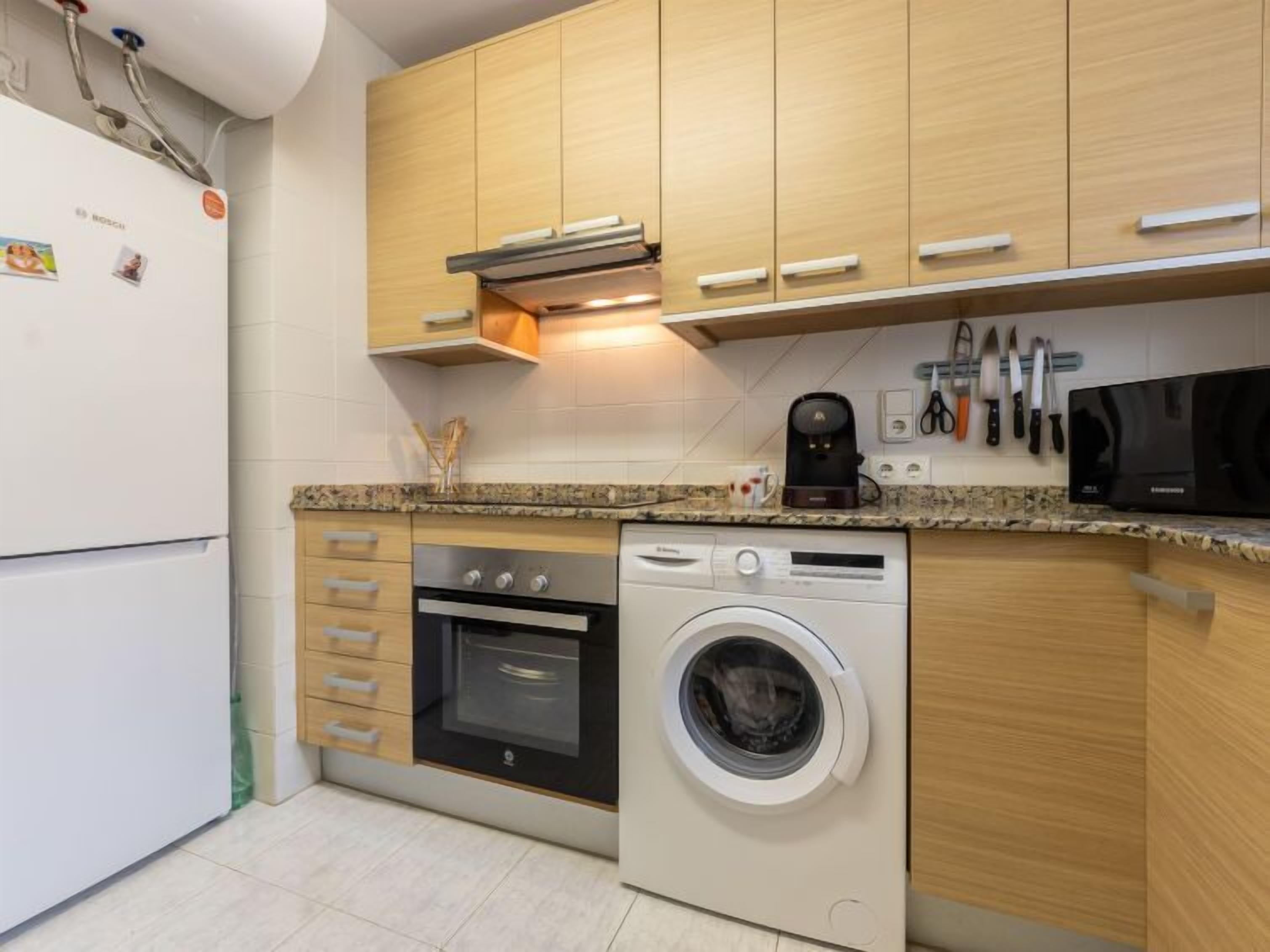 Una cocina compacta con armarios de madera, horno empotrado, lavadora y cafetera.