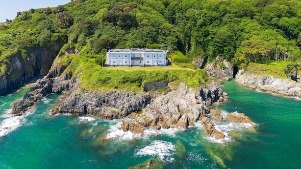 A large, white building with multiple windows, situated on a rocky coastline with lush green hills in the background. - Amazing views -  Woodburner - Wi-Fi (Torpoint)