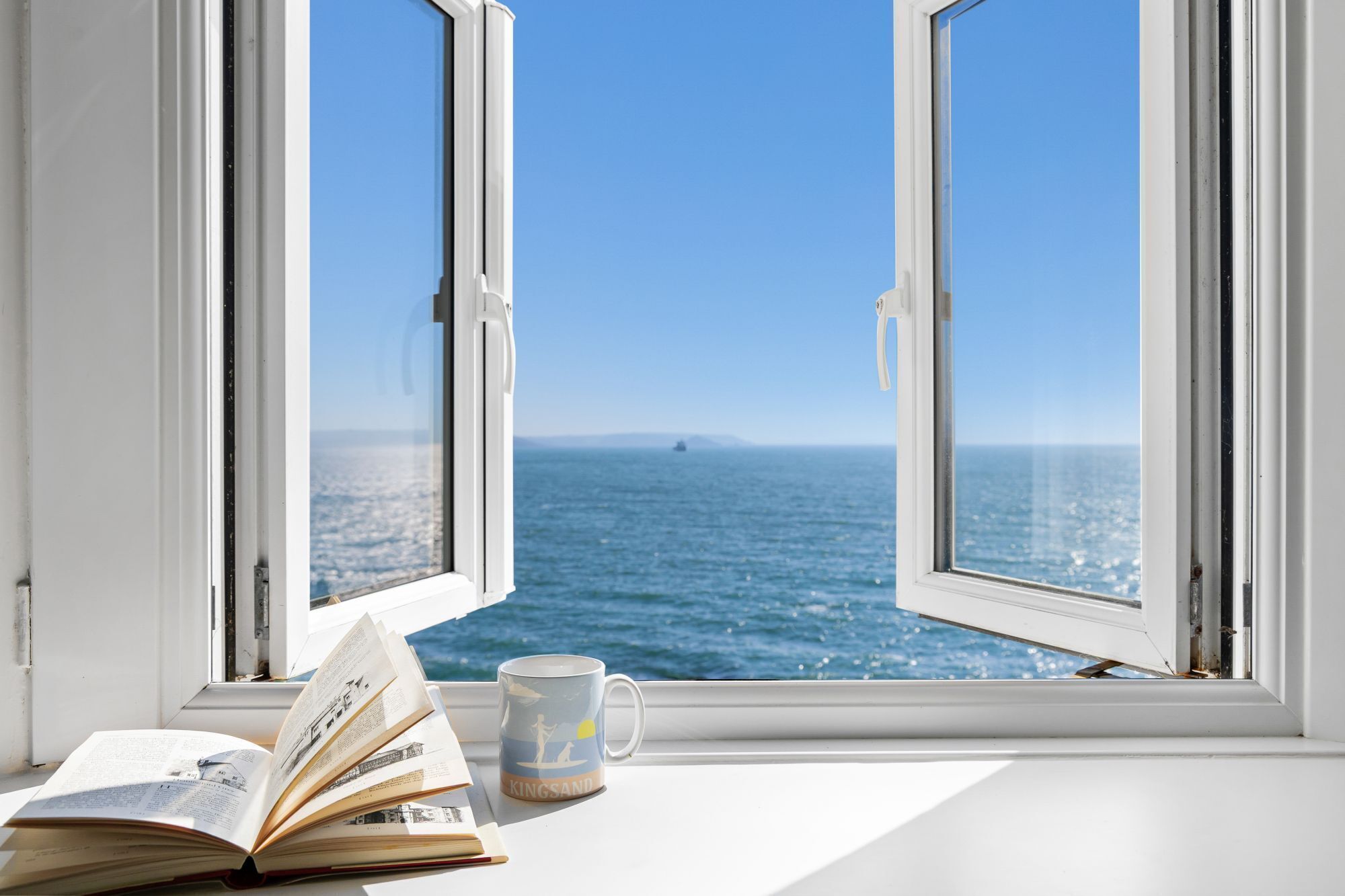 Ein offenes Fenster mit Meerblick, ein Buch und eine Tasse auf dem Fensterbrett.