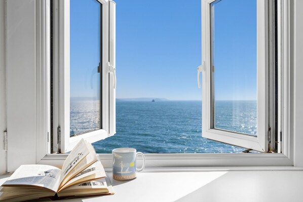 An open window with a view of the sea, a book, and a mug on the windowsill.