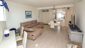 En moderne stue med en beige hjørnesofa, et spisebord med stole, et fjernsyn på et stativ og et køkken i baggrunden.
