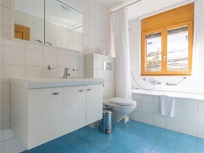 Ein modernes Badezimmer mit einem weißen Waschbecken, Spiegel und WC, Boden in blauem Fliesenmuster und einem Fenster mit Blick ins Freie.
