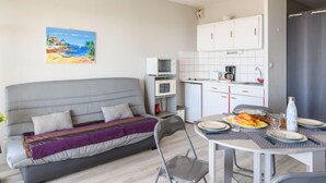 Un espacio habitable compacto con sofá, una mesa de comedor para dos personas, una cocina con armarios blancos y un microondas, y un cuadro de una playa en la pared.