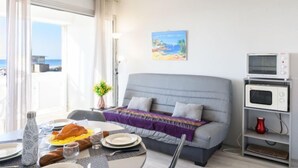 Una habitación moderna con un sofá gris, una mesa de comedor con desayuno, un cuadro y un televisor sobre una estantería.