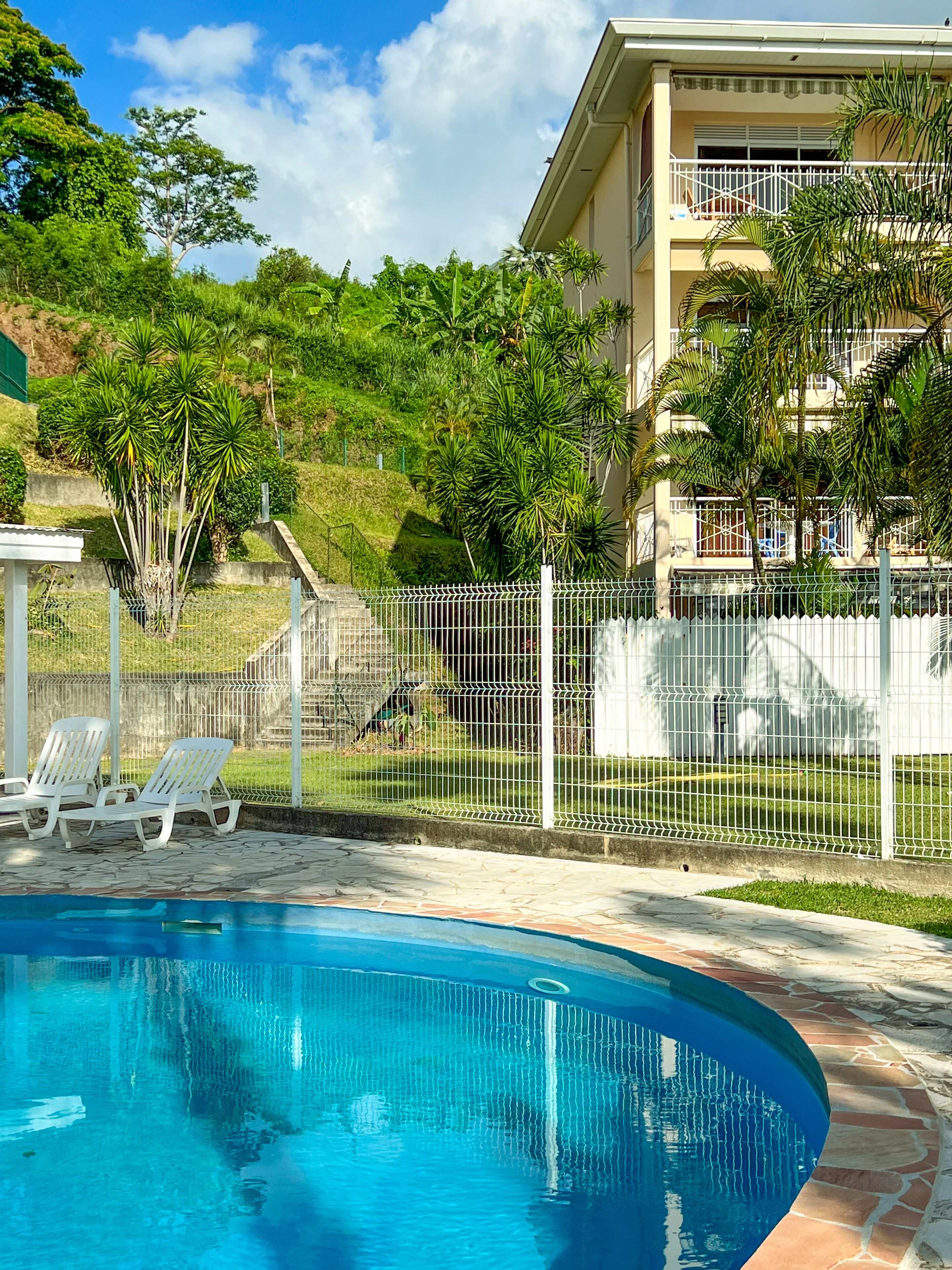 Una piscina con sillones de descanso, un área ajardinada y cercada, y un edificio con balcones.
