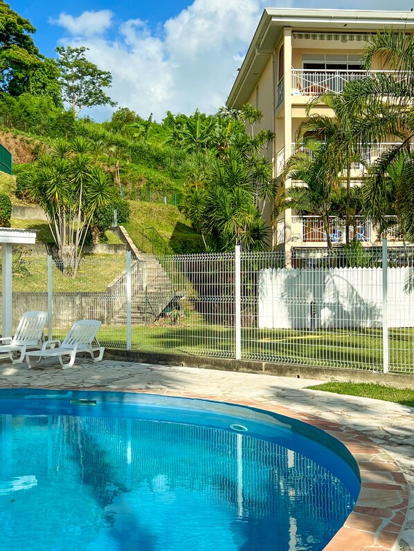 A swimming pool with lounge chairs, a fenced area with greenery, and a building with balconies. - Manui Swimming Pool Condo with Sea & Moorea views (Puna'auia)