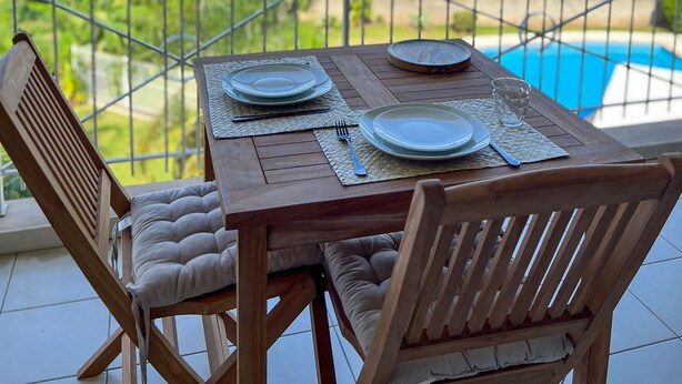 A wooden table set for two with a view of a swimming pool and greenery. - Manui Swimming Pool Condo with Sea & Moorea views (Puna'auia)