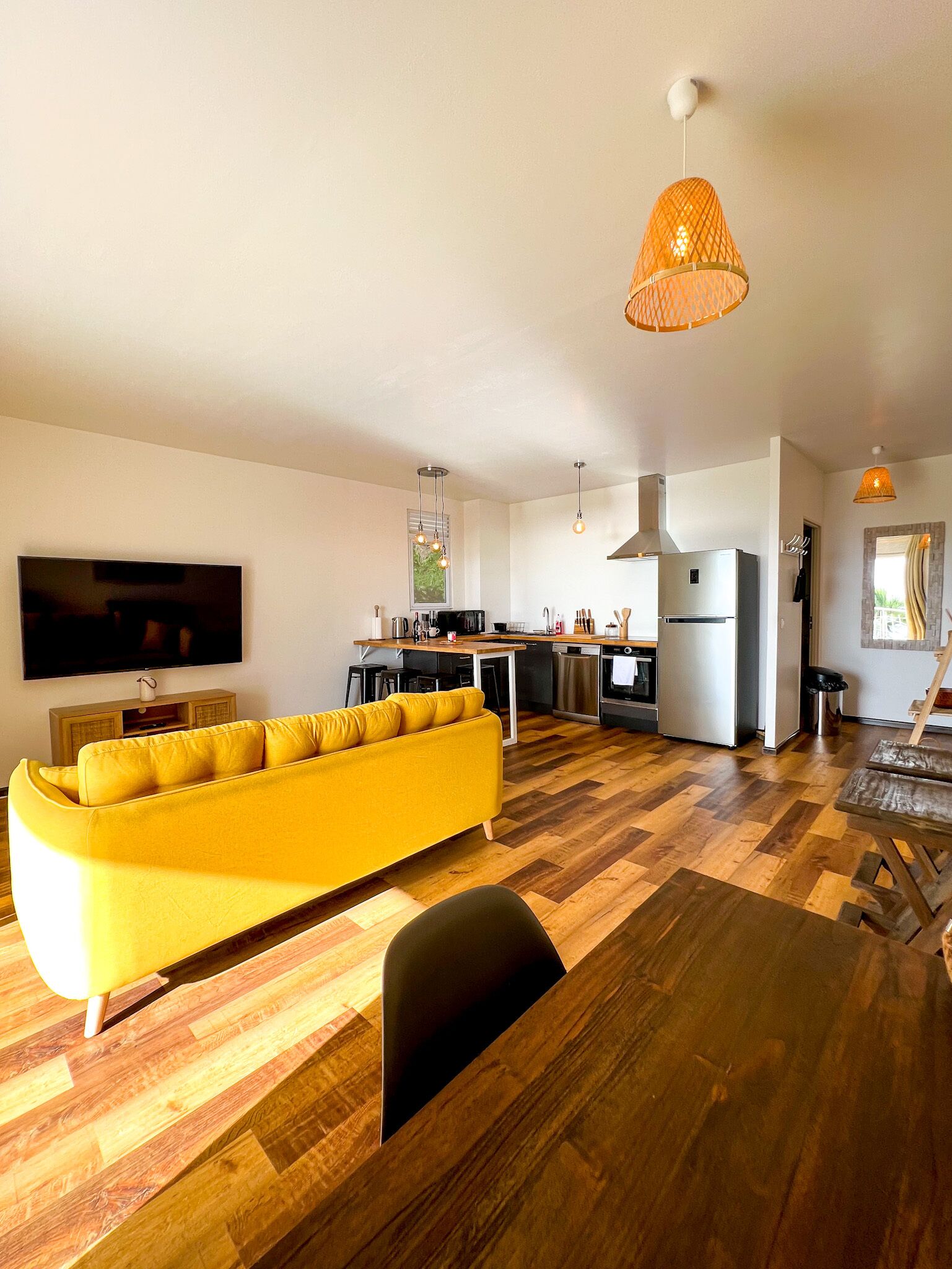 Een moderne woonkamer met een gele bank, houten vloeren, een keukenhoek en hanglampen.