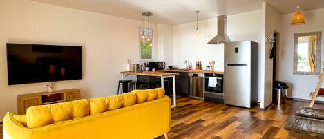 Een moderne woonkamer met een gele bank, houten vloeren, een keukenhoek en hanglampen.