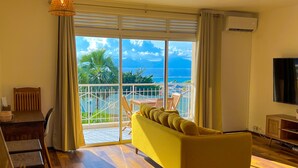 A living room with a yellow sofa, wooden flooring, a dining table with chairs, a hanging lamp, and a view of the ocean through large sliding doors. - Manui Swimming Pool Condo with Sea & Moorea views (Puna'auia)