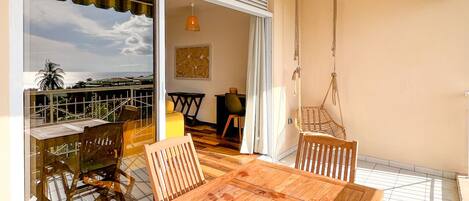 A balcony with a wooden table and chairs, a hanging chair, and a view of the sea.