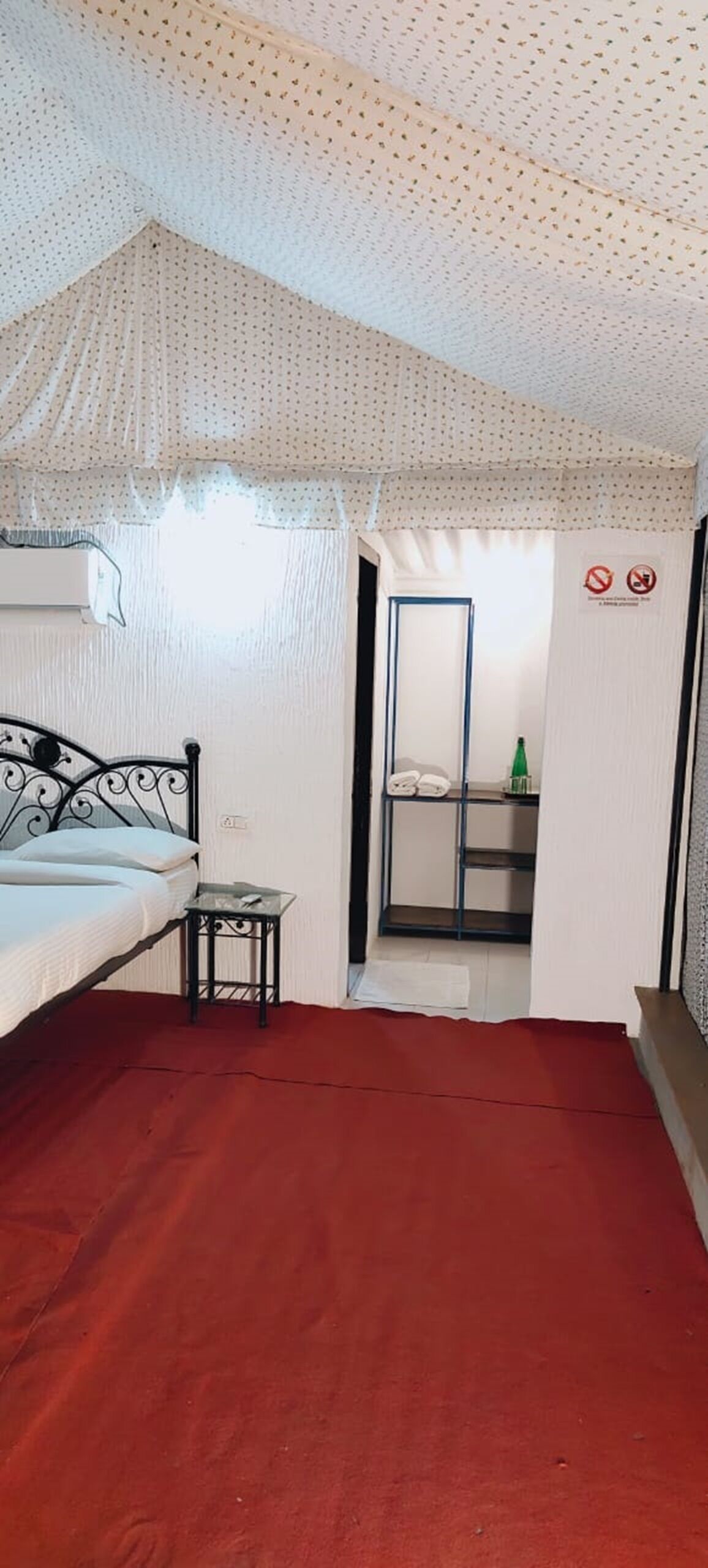 A bedroom with a bed, a small table, a shelf with items, and a red carpet.