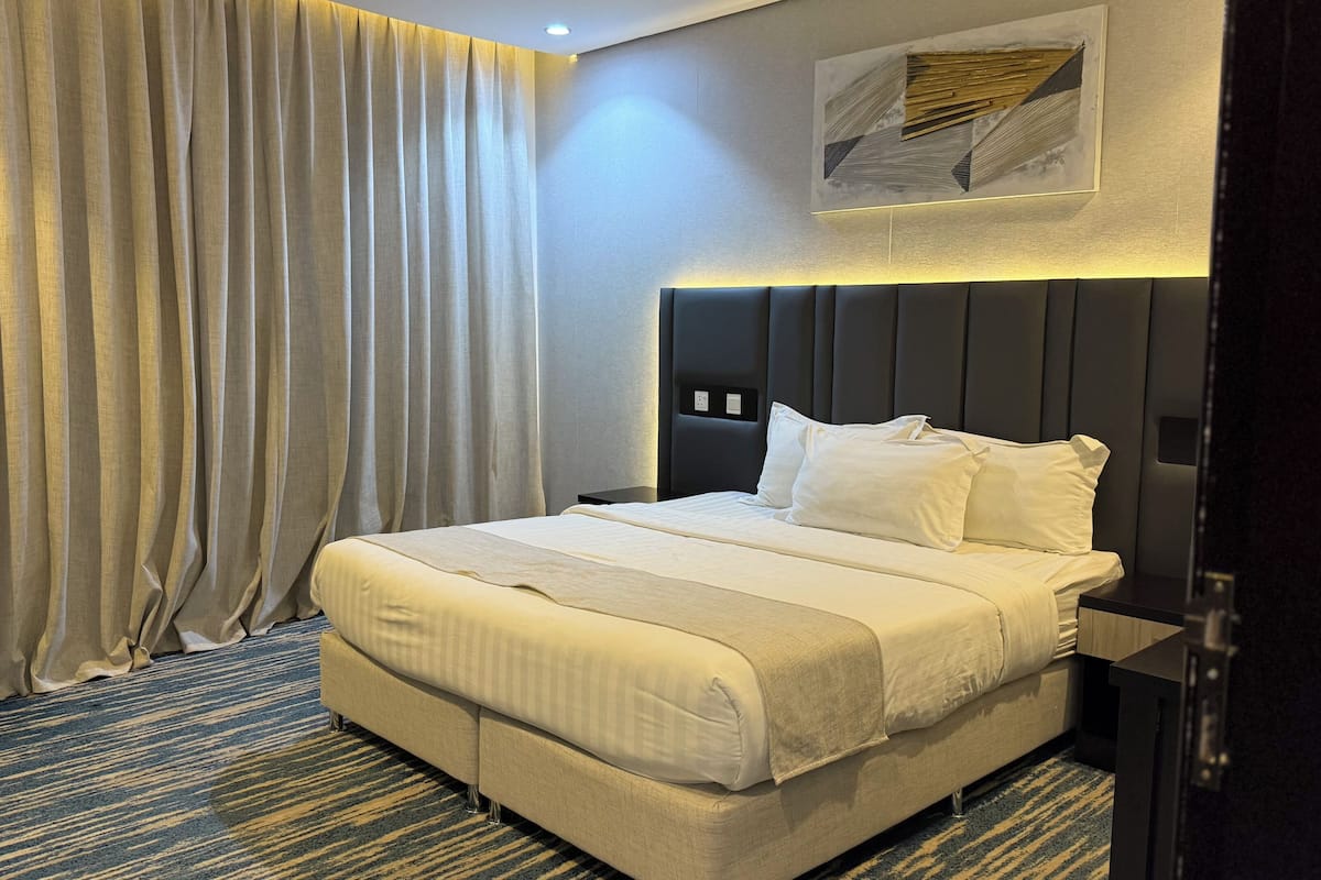 A hotel room with a bed, curtains, a headboard, a wall-mounted artwork, and a carpeted floor.