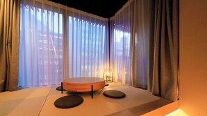 A tatami mat room with a round table and two cushions, large windows with sheer curtains, and a floor lamp. - AMANECER HOTEL (Tokyo)
