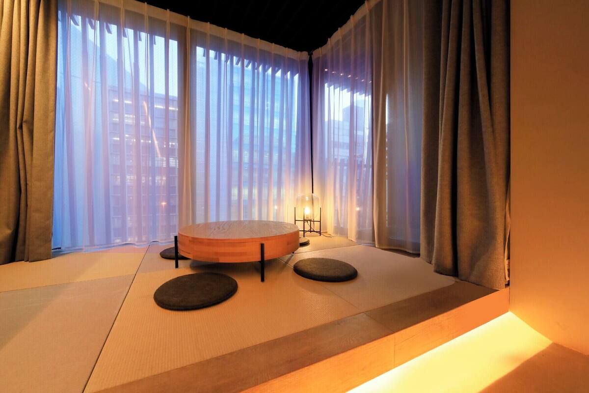 A tatami mat room with a round table and two cushions, large windows with sheer curtains, and a floor lamp.