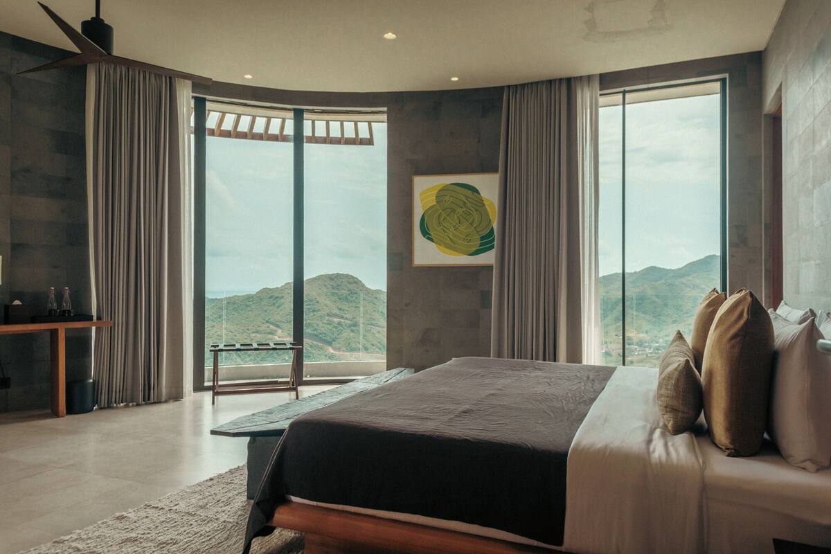 A bedroom with a large bed, a bench, and a view of mountains through large windows.