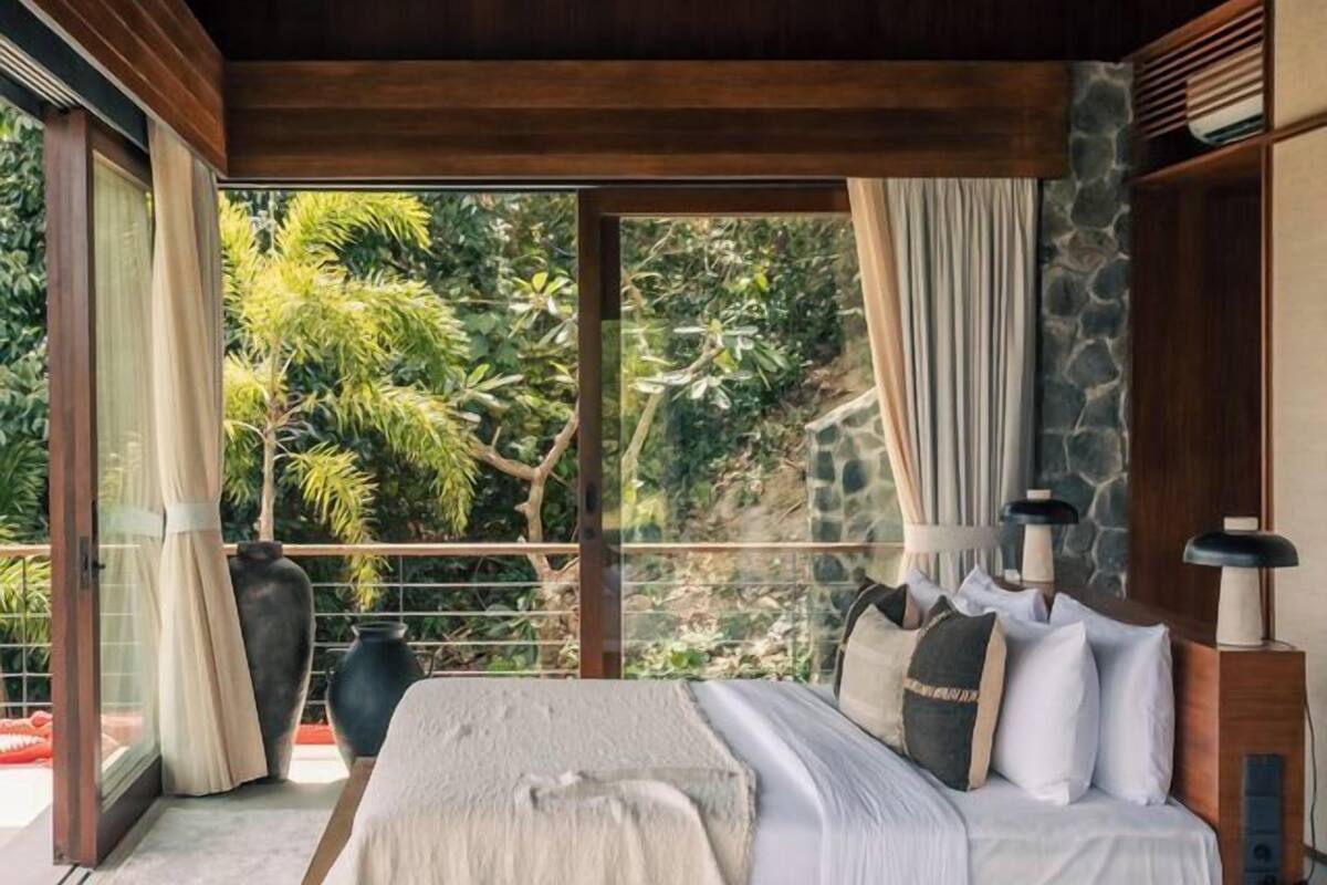 A bedroom with a large bed, wooden ceiling, stone wall, and a view of greenery outside.