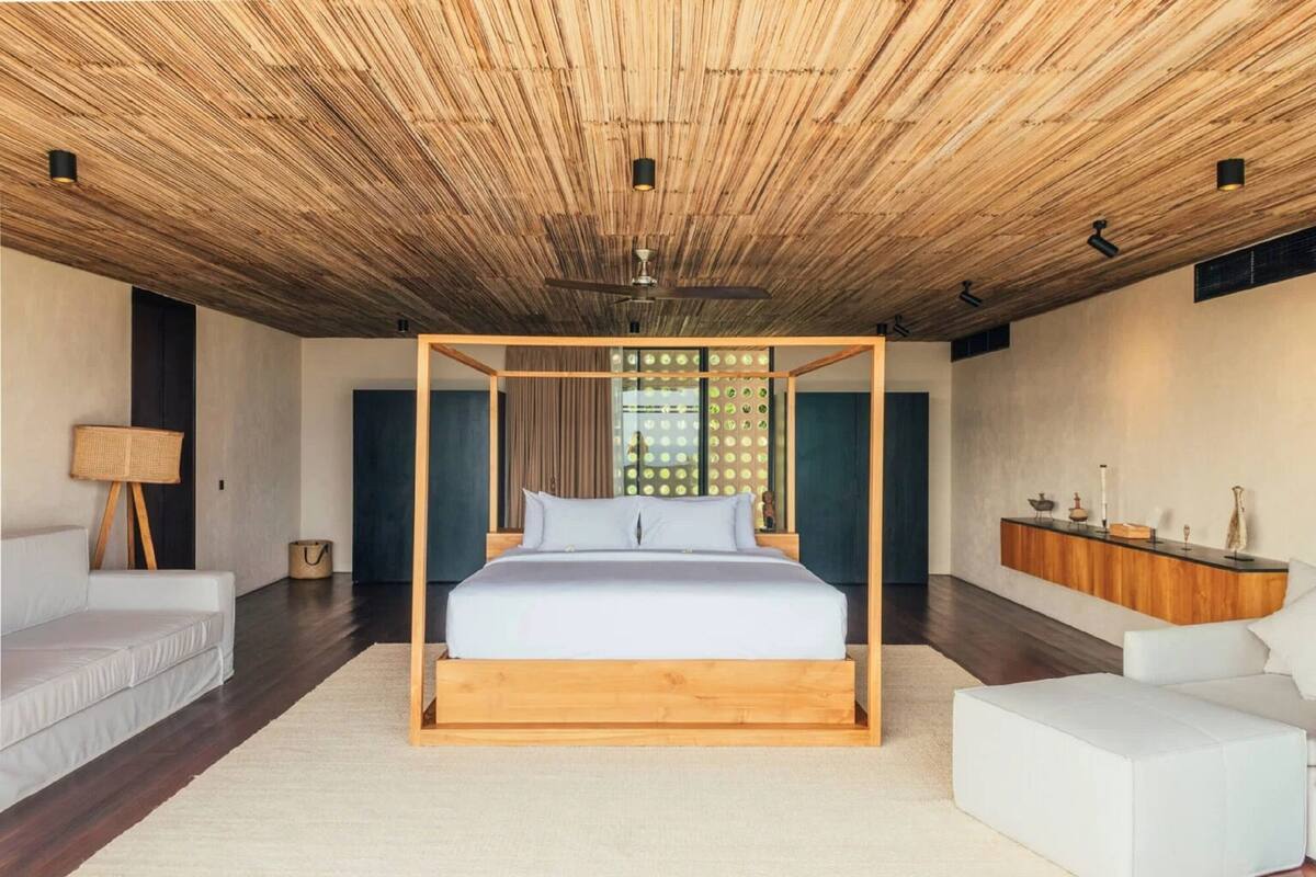 A modern bedroom with a wooden ceiling, a central bed with a wooden frame, a white sofa, a side table, and a cabinet with decorative items.