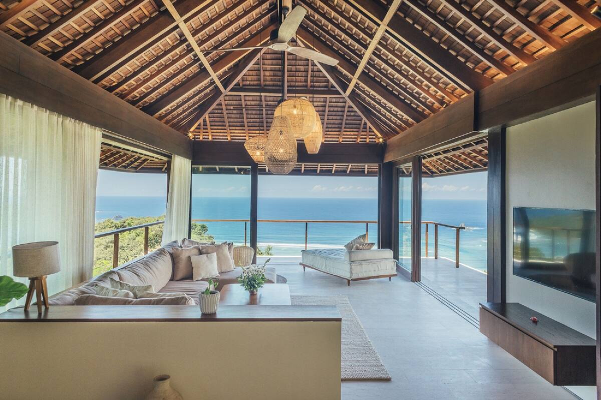 A spacious living area with a view of the ocean, a ceiling fan, and a large TV screen.