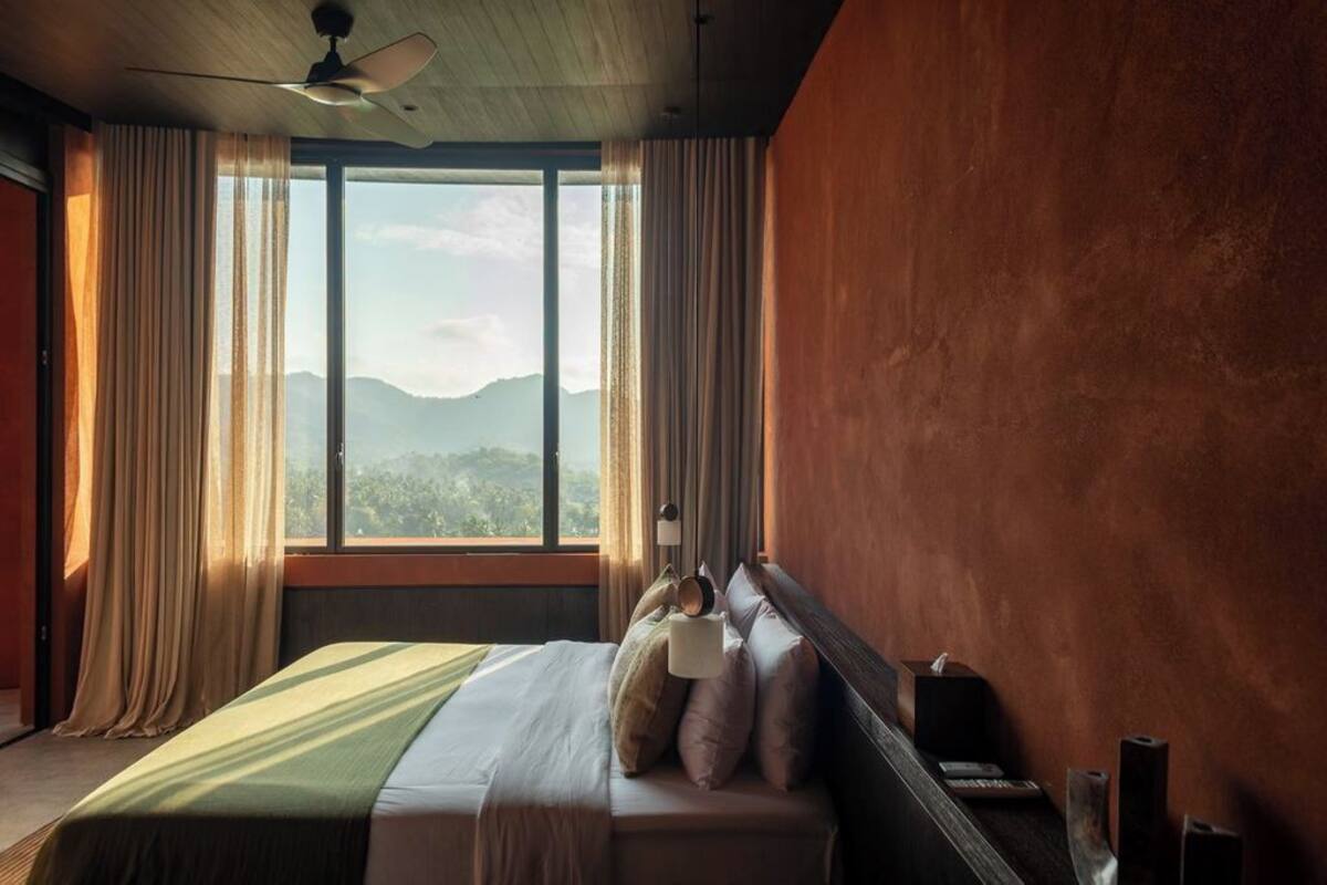 A bedroom with a bed, curtains, a nightstand, a lamp, and a view of mountains through large windows.
