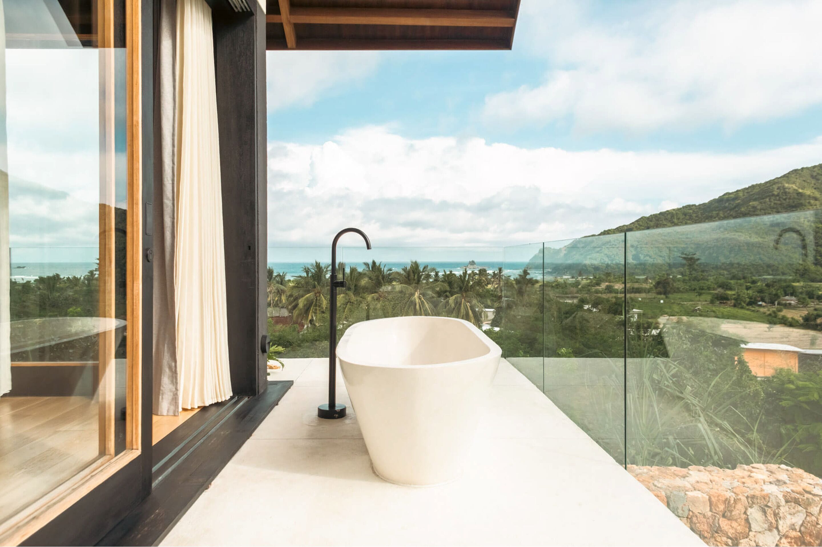 A modern bathroom with a large white bathtub, a glass-enclosed shower area, and a view of a coastal landscape.