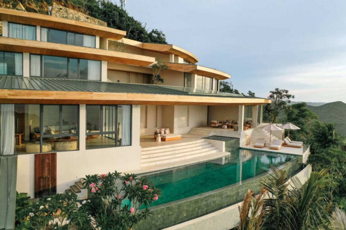 A modern multi-story house with a large swimming pool, outdoor seating, and a view of the hills.
