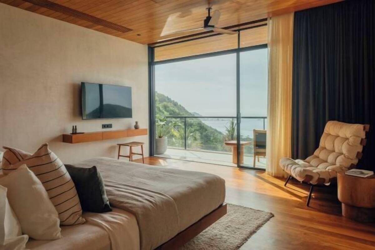 A modern bedroom with a large bed, a TV mounted on the wall, a chair, and a view of the outdoors.