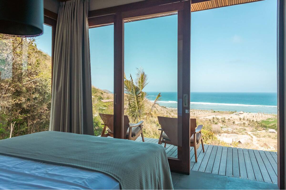 A bedroom with a large bed, wooden chairs, and a view of the ocean.