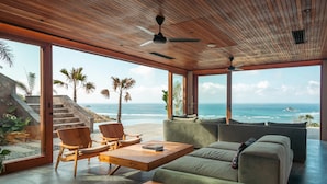 A modern living room with a wooden ceiling, large windows, a green sofa, wooden chairs, and a small table. - Tampah Hills (Kuta)