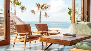 A room with two wooden chairs, a wooden table, and a green sofa. There is a view of the ocean and palm trees outside. - Tampah Hills (Kuta)
