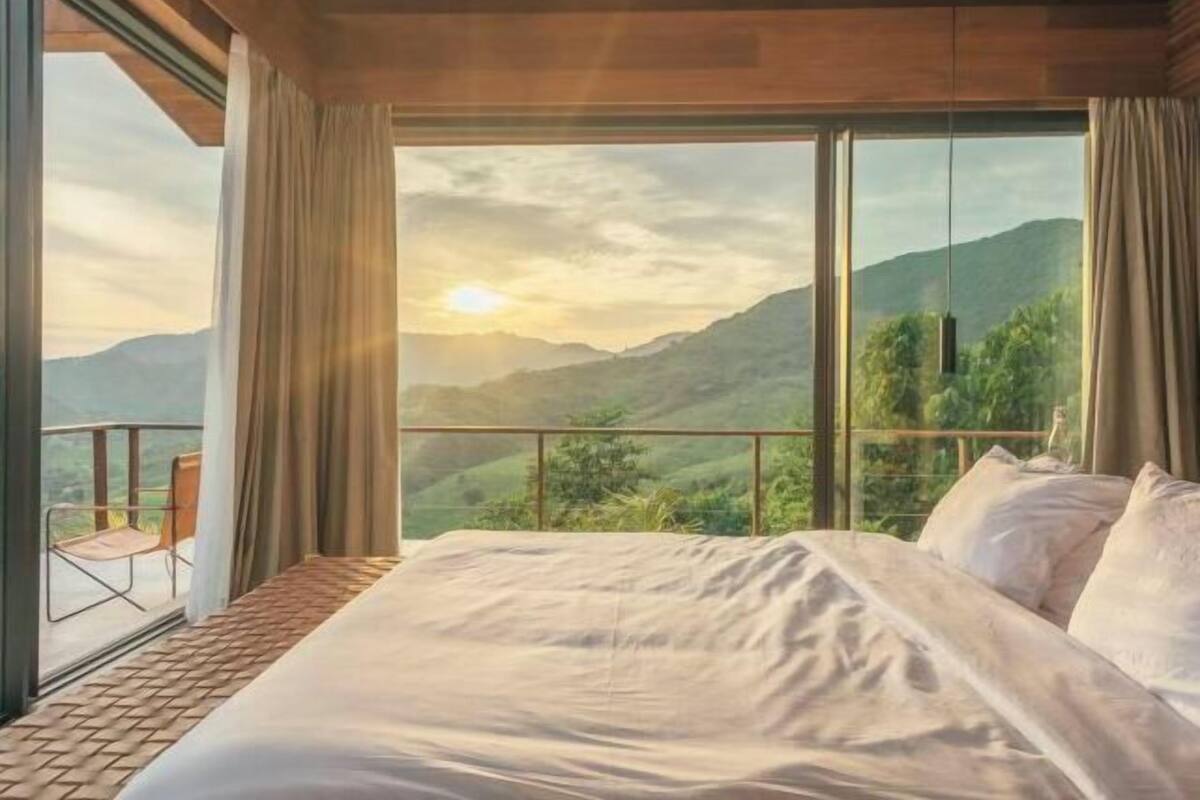 A bedroom with a large bed, a view of mountains, and a balcony with a chair.
