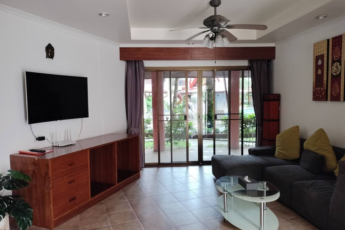 A living room with a flat-screen TV, wooden entertainment unit, glass coffee table, and a dark sofa with cushions.